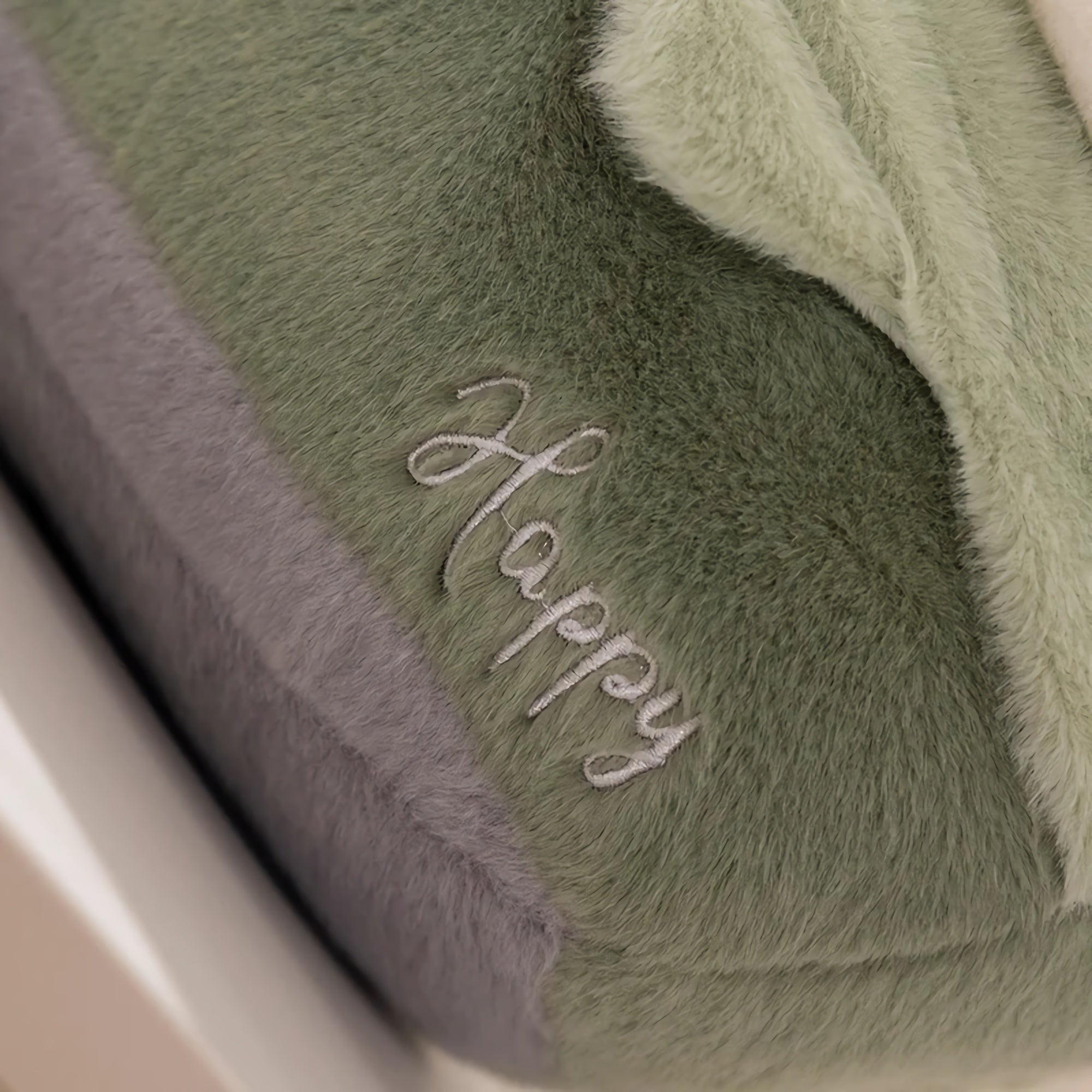 Close-up of a plush pillow with the word "Happy" embroidered in silver on a soft green and gray fabric.