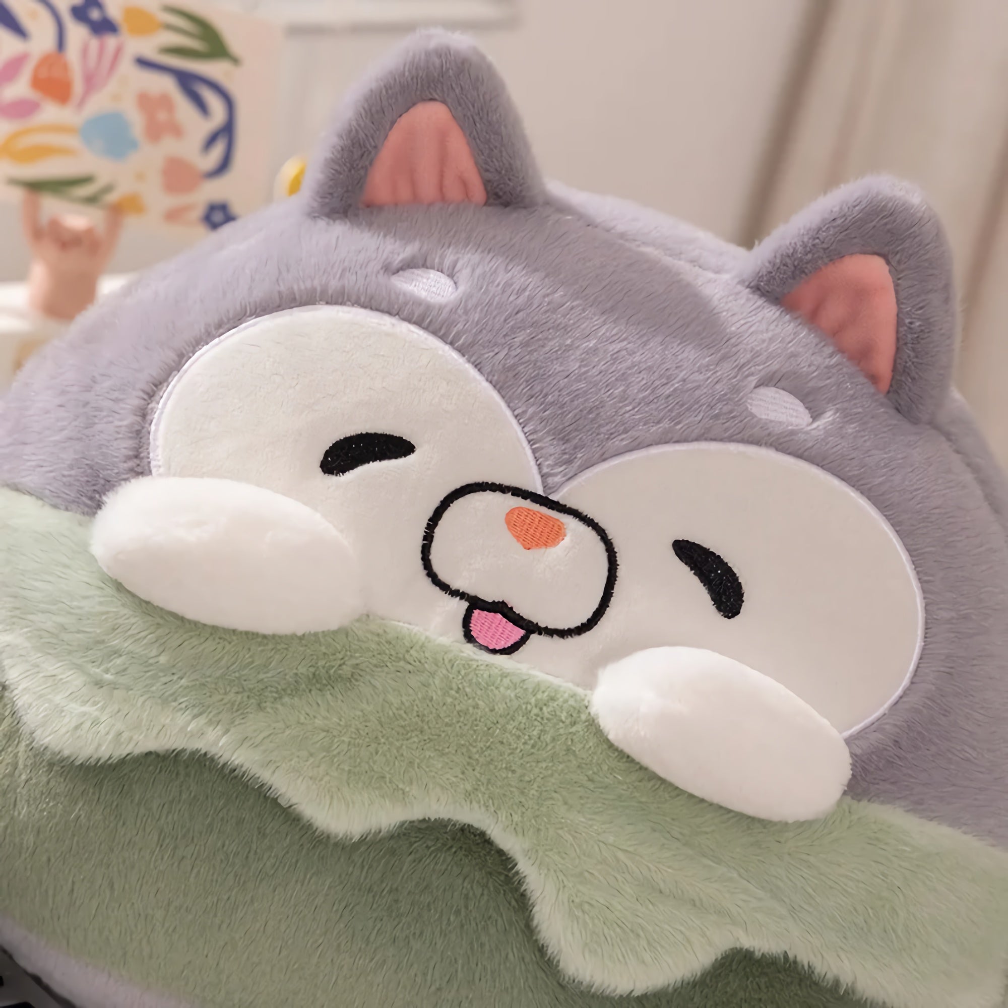 Plush pillow with a cute animal face, gray ears, and a green blanket.