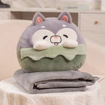 A plush pillow shaped like a smiling animal sits on top of a folded gray travel blanket.