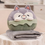 A plush pillow shaped like a smiling animal sits on top of a folded gray travel blanket.