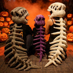 Three people sit with dinosaur skeleton plush hoods and spines, surrounded by carved pumpkins and fog.