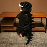 Child wearing a dinosaur skeleton plush hood and spine, sitting at a wooden table with bare feet.
