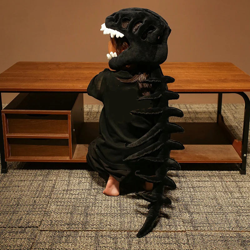 Child wearing a dinosaur skeleton plush hood and spine, sitting at a wooden table with bare feet.