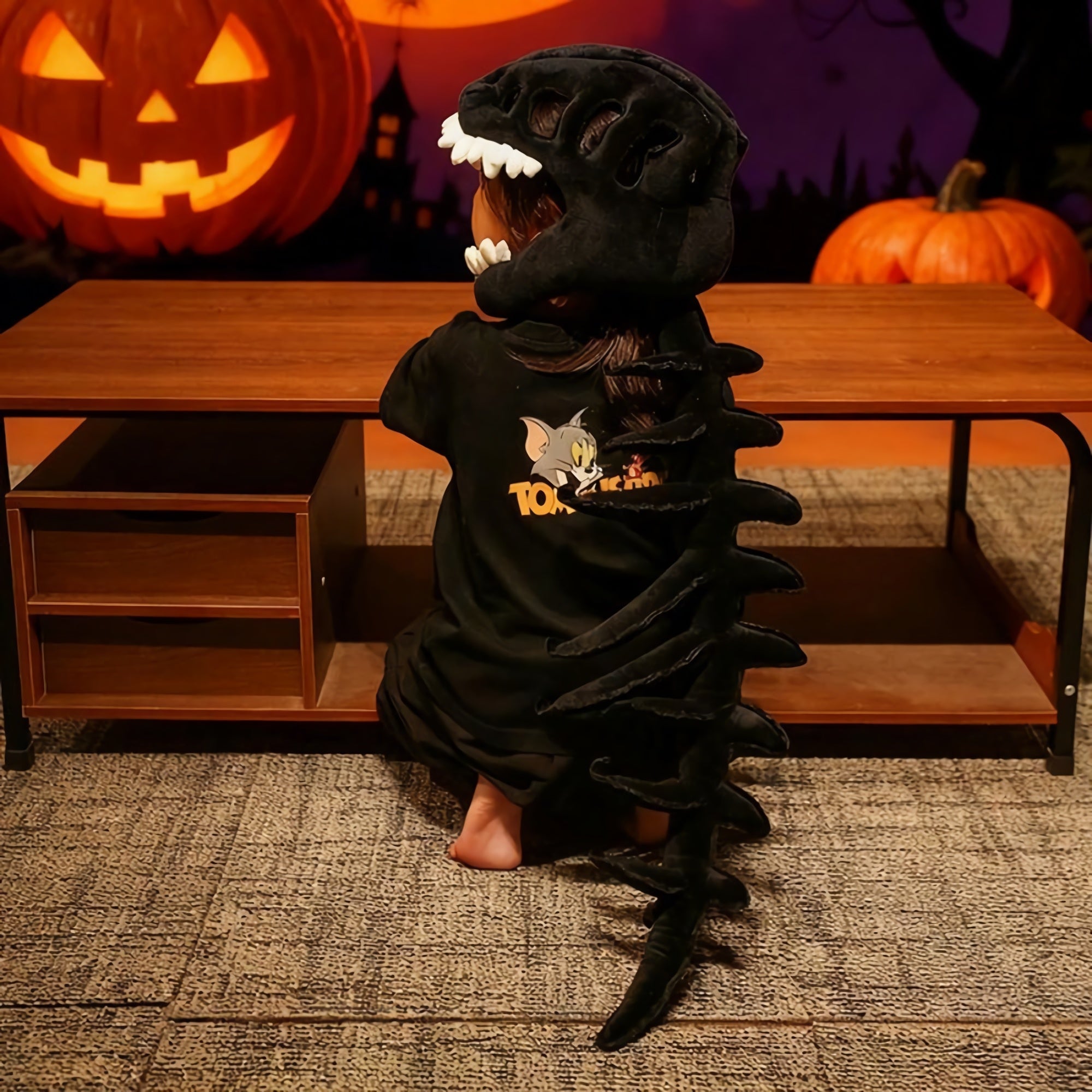 Child wearing a dinosaur skeleton plush hood and spine, sitting at a table with pumpkins in the background.