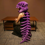 A person wearing a purple dinosaur skeleton plush hood and spine sits at a table, facing away from the camera.