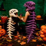 A child and adult wear dinosaur skeleton plush hoods and spines, sitting at a table with pumpkins and autumn leaves.