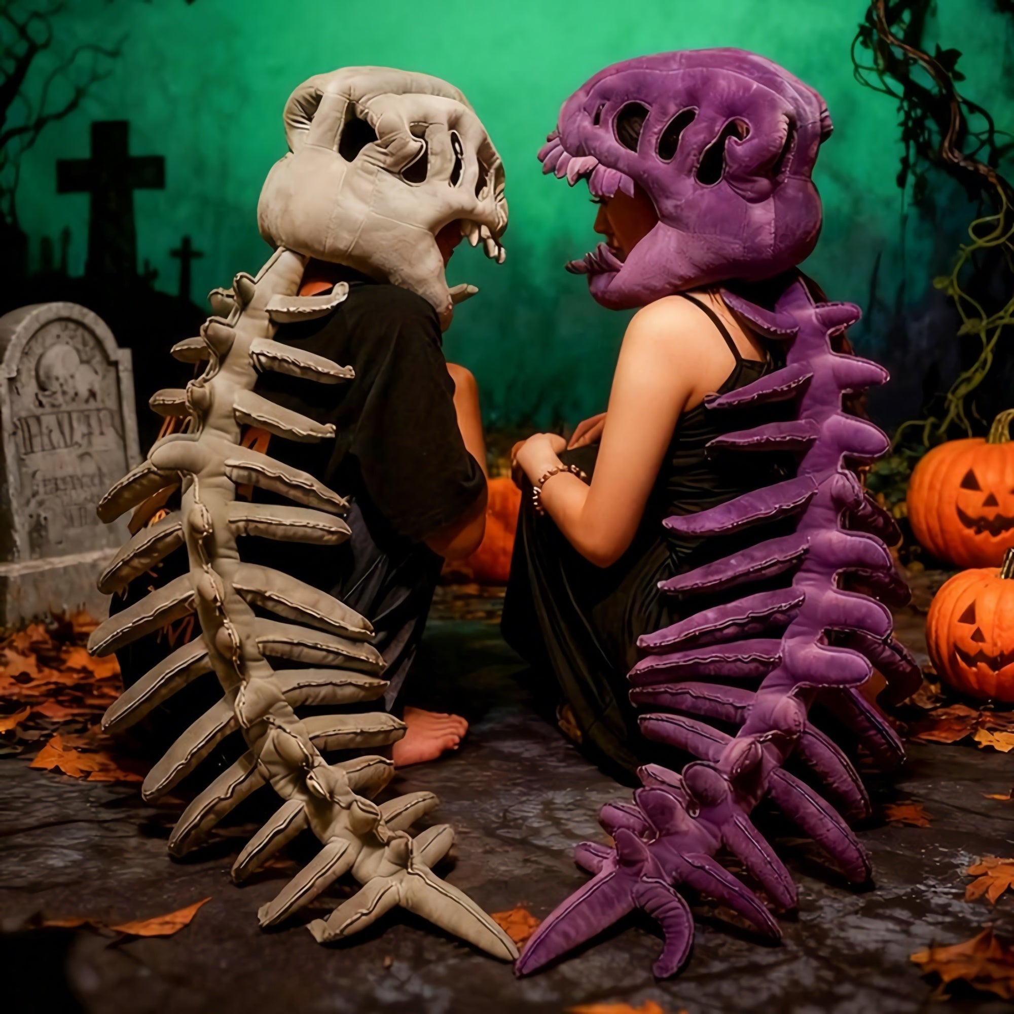 Two people sit back-to-back wearing dinosaur skeleton plush hoods and spines, surrounded by pumpkins and a graveyard backdrop
