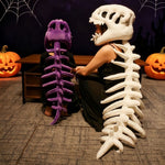 Two people sit with dinosaur skeleton plush hoods and spines, one in purple and the other in white, near Halloween decoration