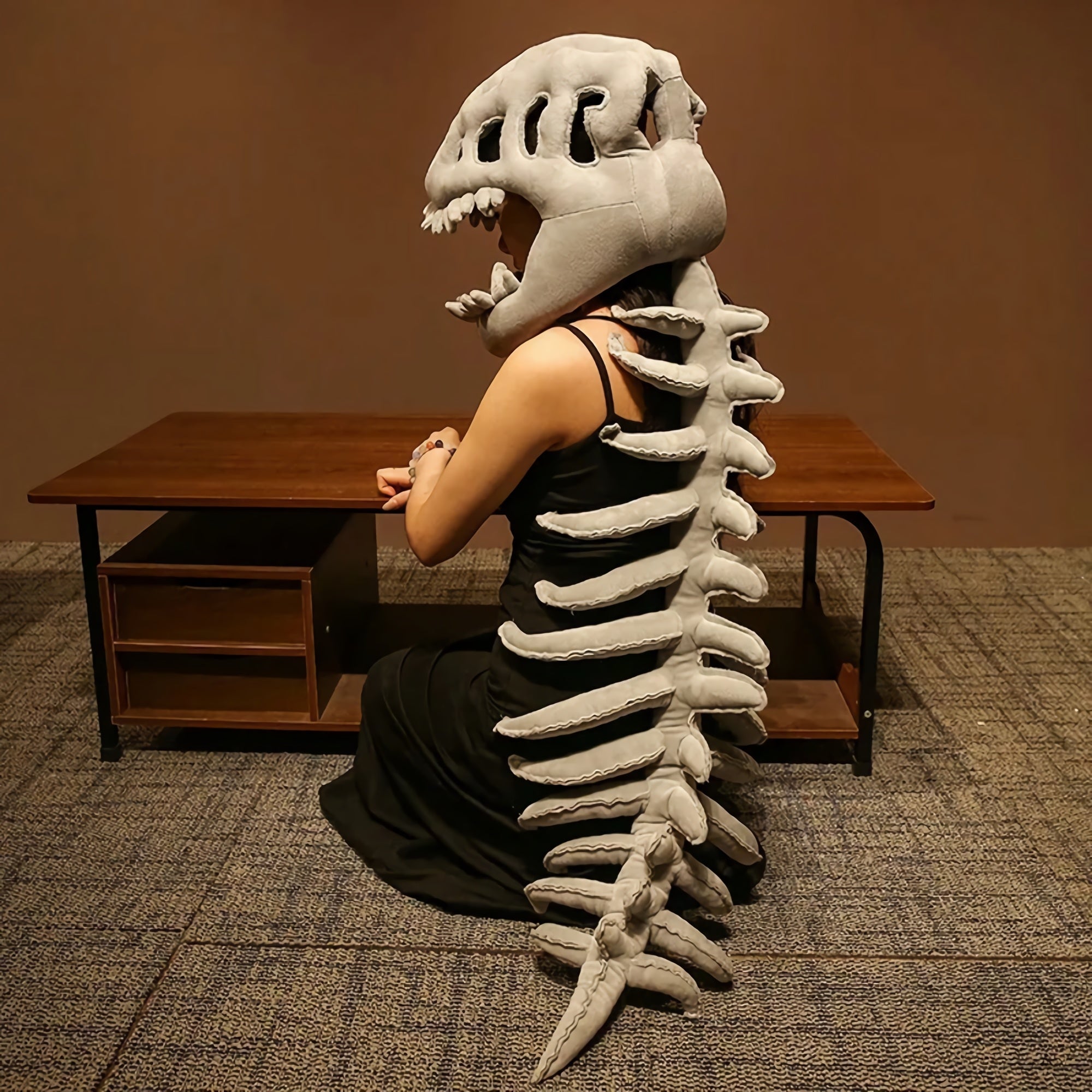 A person wearing a dinosaur skeleton plush hood and spine sits at a table, facing away from the camera.