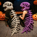 Two people sit back-to-back wearing dinosaur skeleton plush hoods and spines, one gray and one purple, with pumpkins nearby.