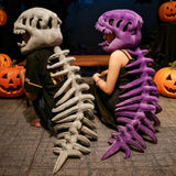 Two people sit back-to-back wearing dinosaur skeleton plush hoods and spines, one gray and one purple, with pumpkins nearby.