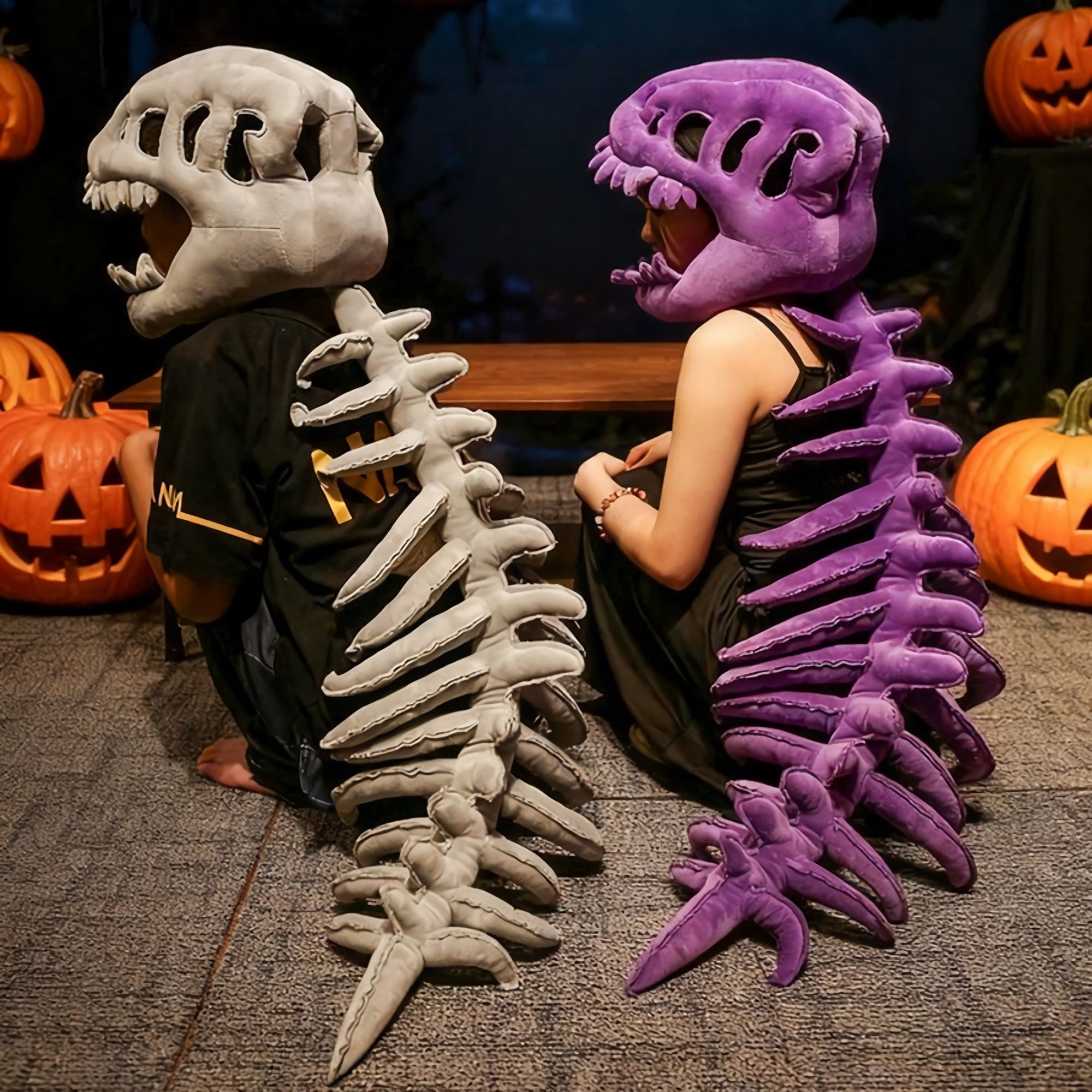 Two people sit back-to-back wearing dinosaur skeleton plush hoods and spines, one gray and one purple, with pumpkins nearby.