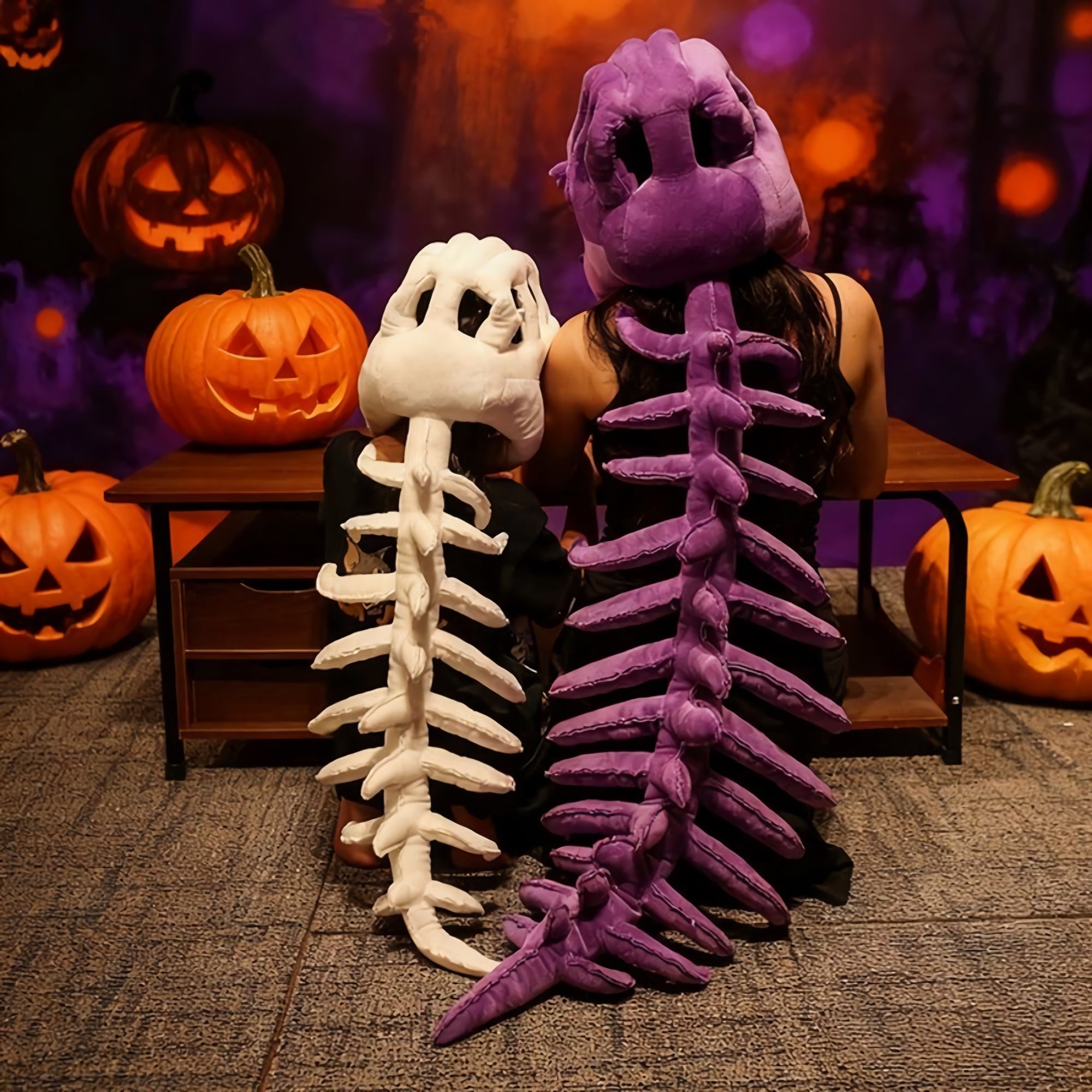 A child and an adult wear dinosaur skeleton plush hoods and spines, sitting in front of carved pumpkins.