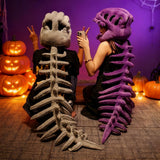 Two people wearing dinosaur skeleton plush hoods and spines, sitting back-to-back with pumpkins in the background.