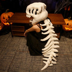 A person wearing a dinosaur skeleton plush hood and spine, seated near carved pumpkins.
