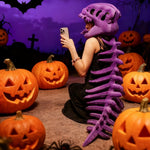 A person wearing a purple dinosaur skeleton plush hood and spine sits among carved pumpkins, looking at a phone.