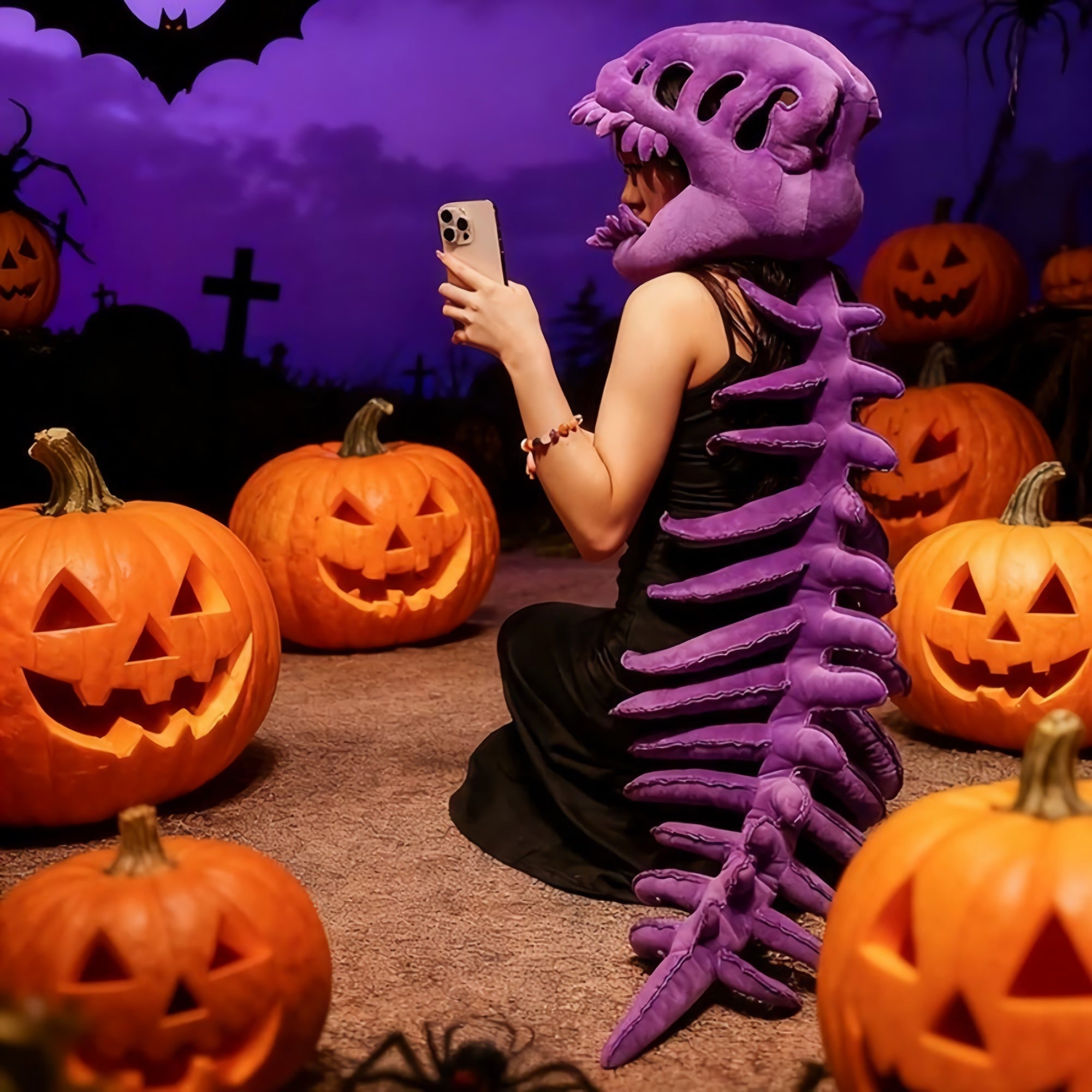 A person wearing a purple dinosaur skeleton plush hood and spine sits among carved pumpkins, looking at a phone.