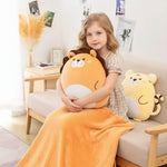 A girl sits on a couch holding a plush lion pillow, with a yellow tiger pillow beside her and a blanket draped over her legs.