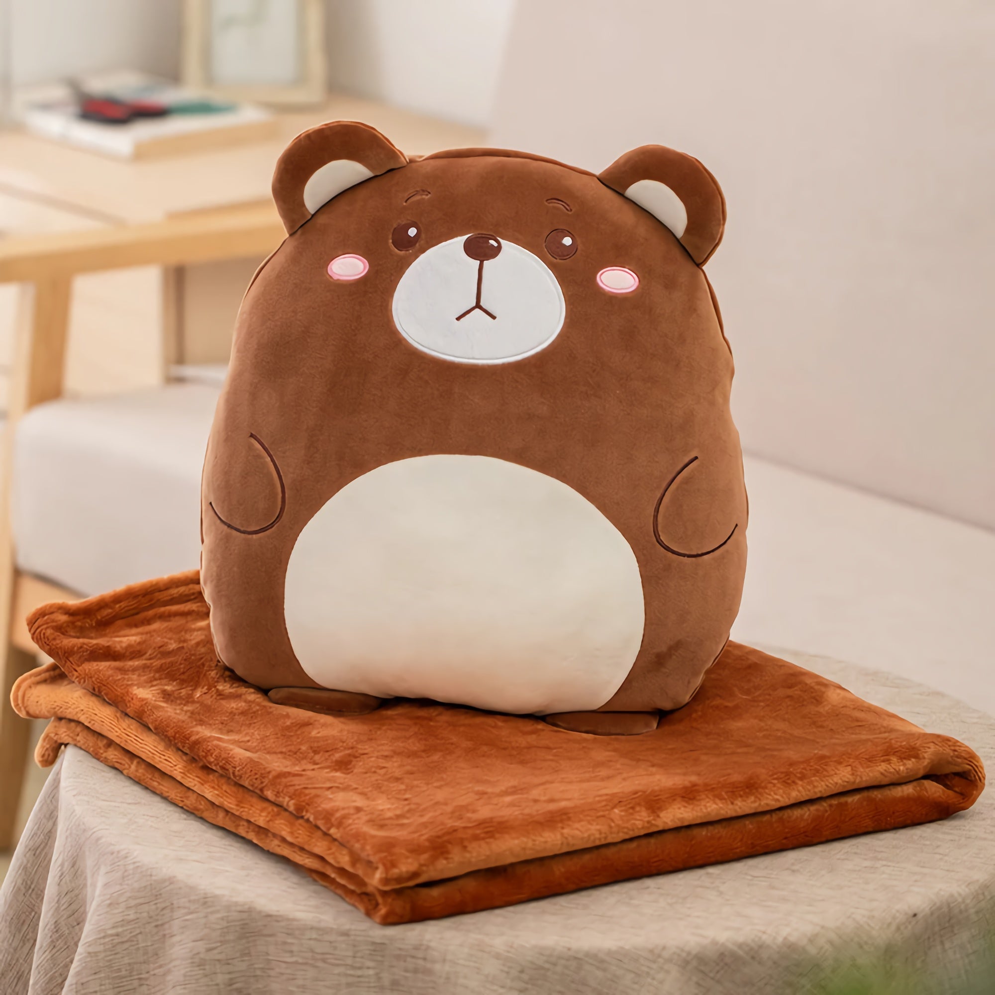 A plush bear pillow sits on top of a folded orange travel blanket on a table.