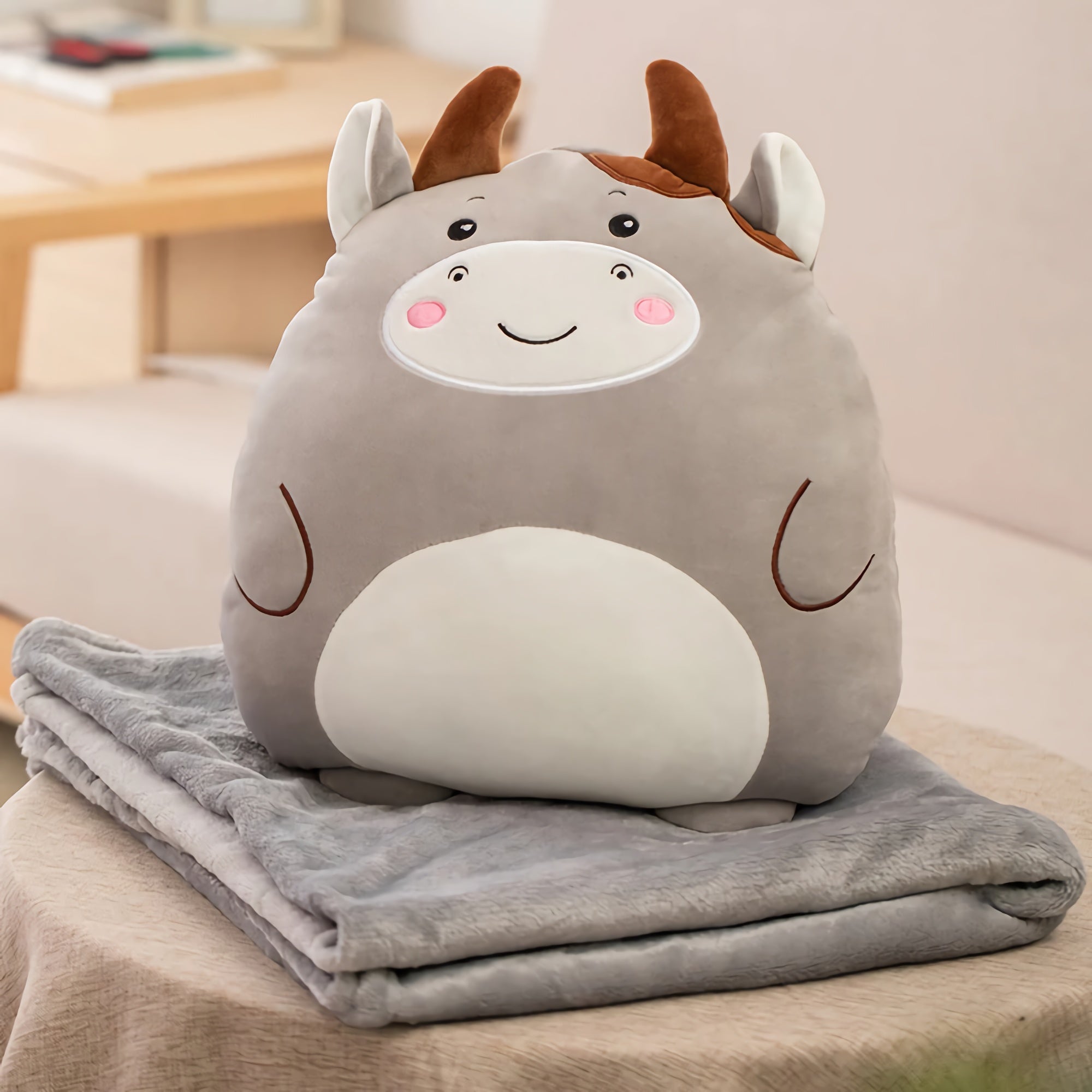 A plush pillow shaped like a smiling gray animal sits on a folded gray blanket on a table.