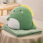 A green plush dinosaur pillow sits on a folded soft blanket on a table.