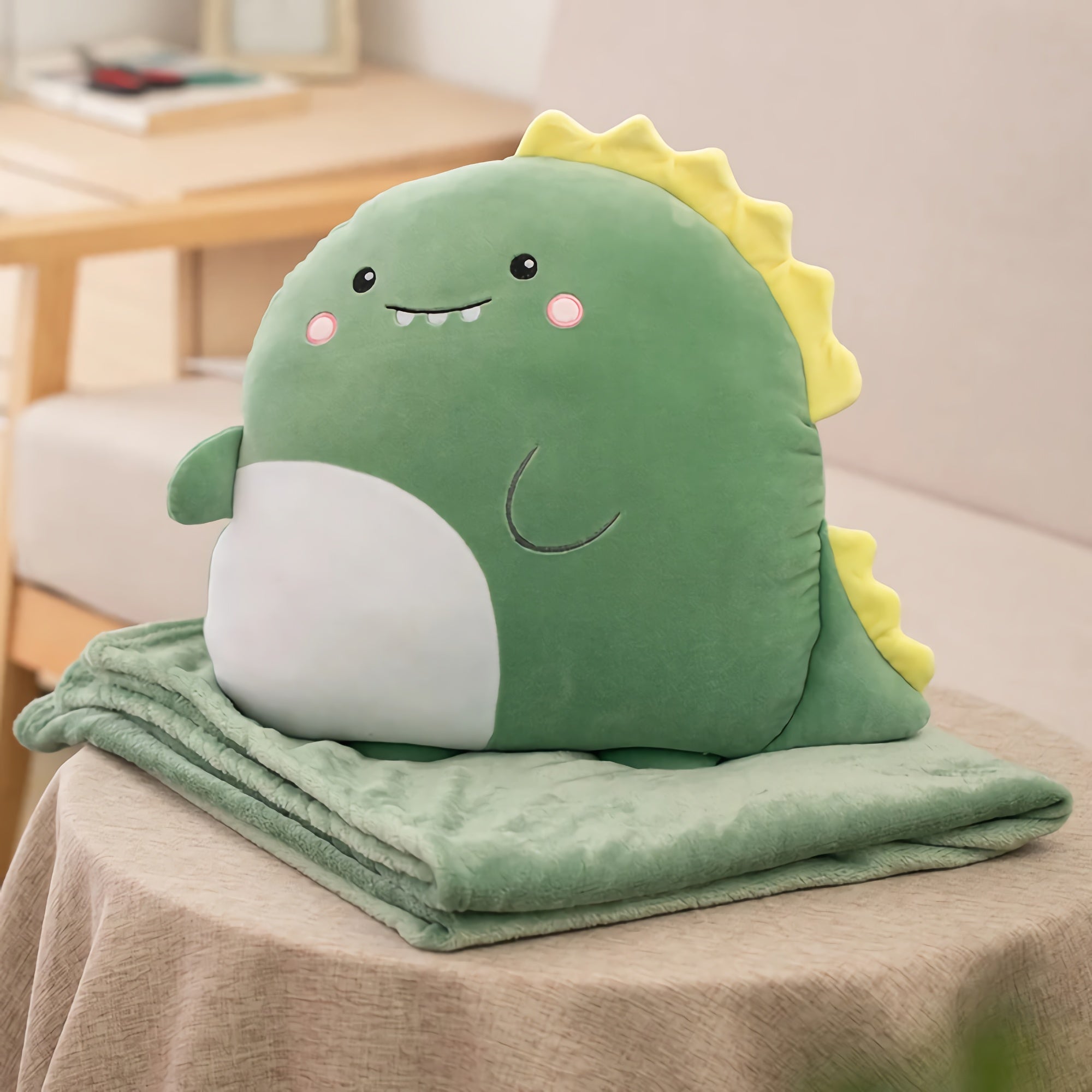 A green plush dinosaur pillow sits on a folded soft blanket on a table.