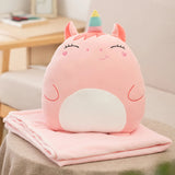 A pink plush pillow shaped like a unicorn rests on a folded pink travel blanket on a table.