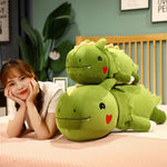 A woman rests her chin on her hands beside two large green dinosaur plush pillows on a bed.
