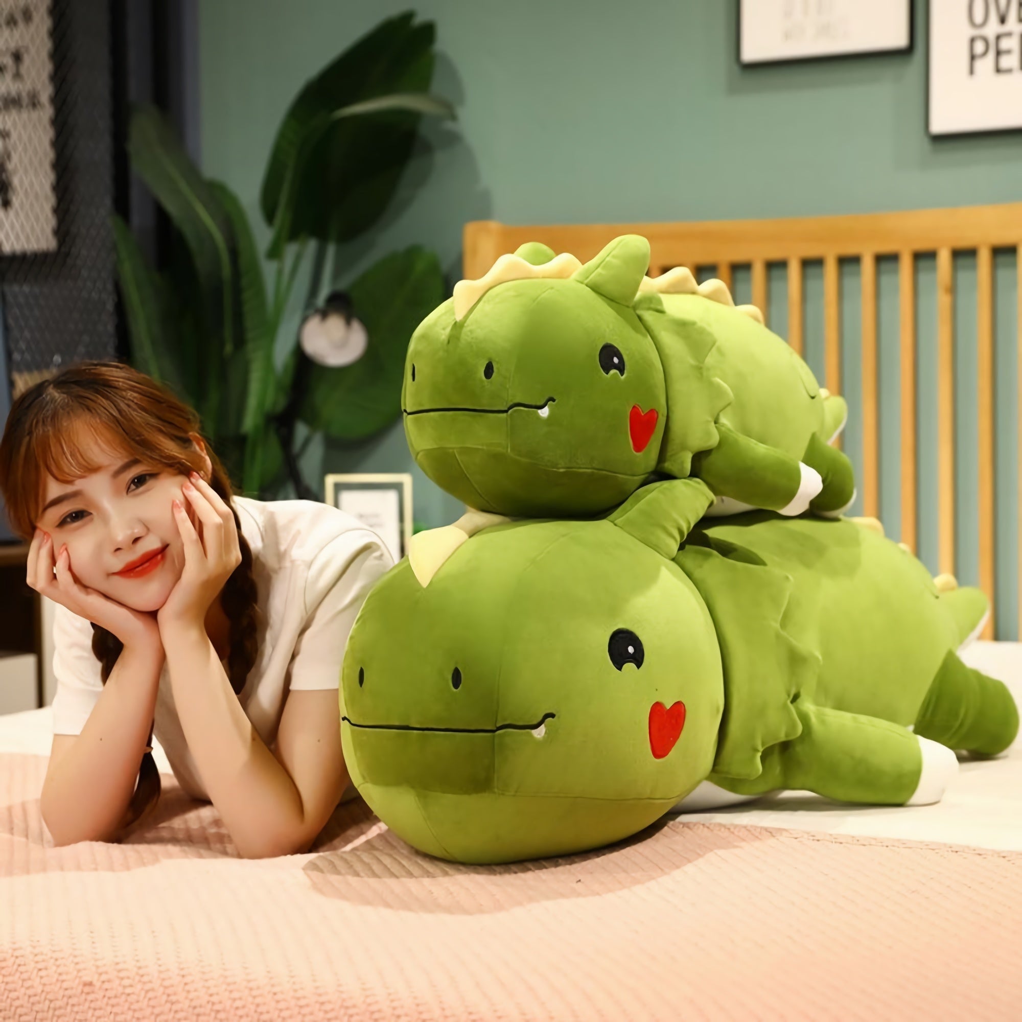 A woman rests her chin on her hands beside two large green dinosaur plush pillows on a bed.