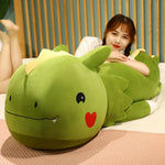 A green dinosaur plush pillow with a smiling face, lying on a bed beside a person.