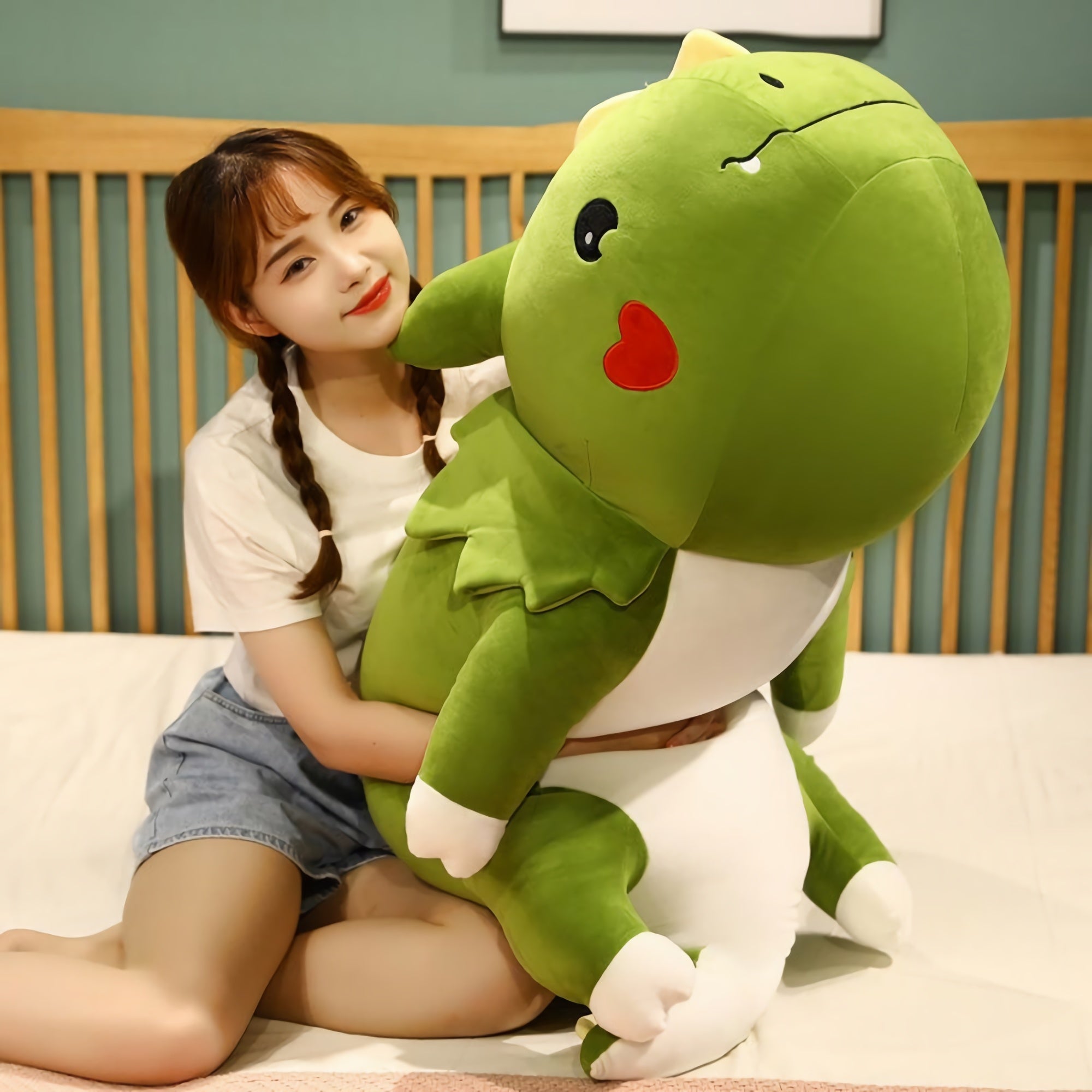 A person sits on a bed, hugging a large green dinosaur plush pillow with a smiling face.