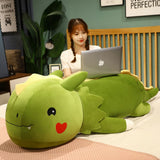 A person sits on a bed using a laptop, with a large green dinosaur plush pillow beside them.
