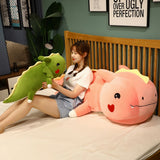 A girl sits on a bed holding a small green dinosaur plush while a large pink dinosaur plush rests beside her.