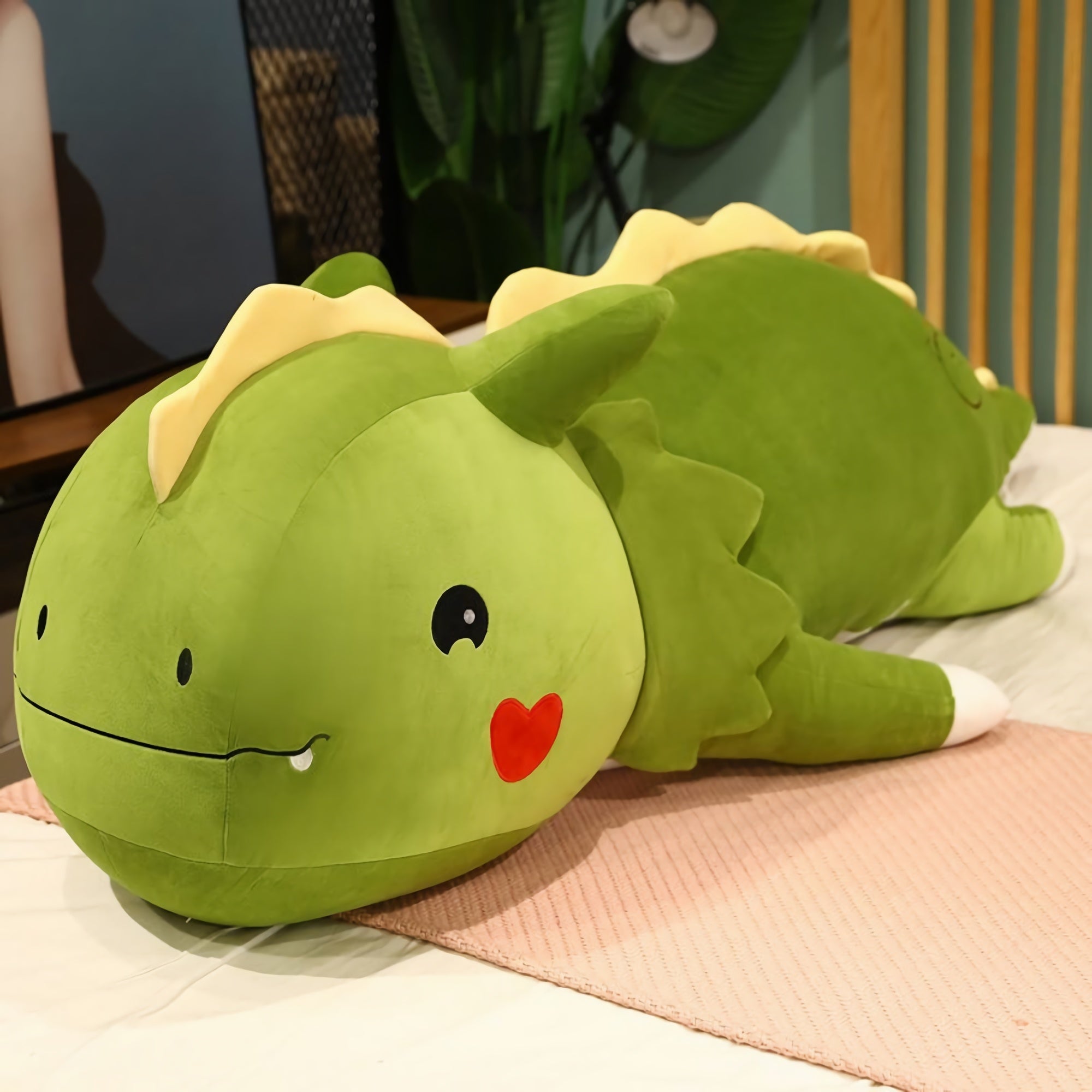 Green dinosaur plush pillow with a smiling face and small heart, lying on a bed with a pink mat.
