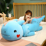 A person lies on a bed next to a large blue dinosaur plush pillow with a smiling face and a heart.