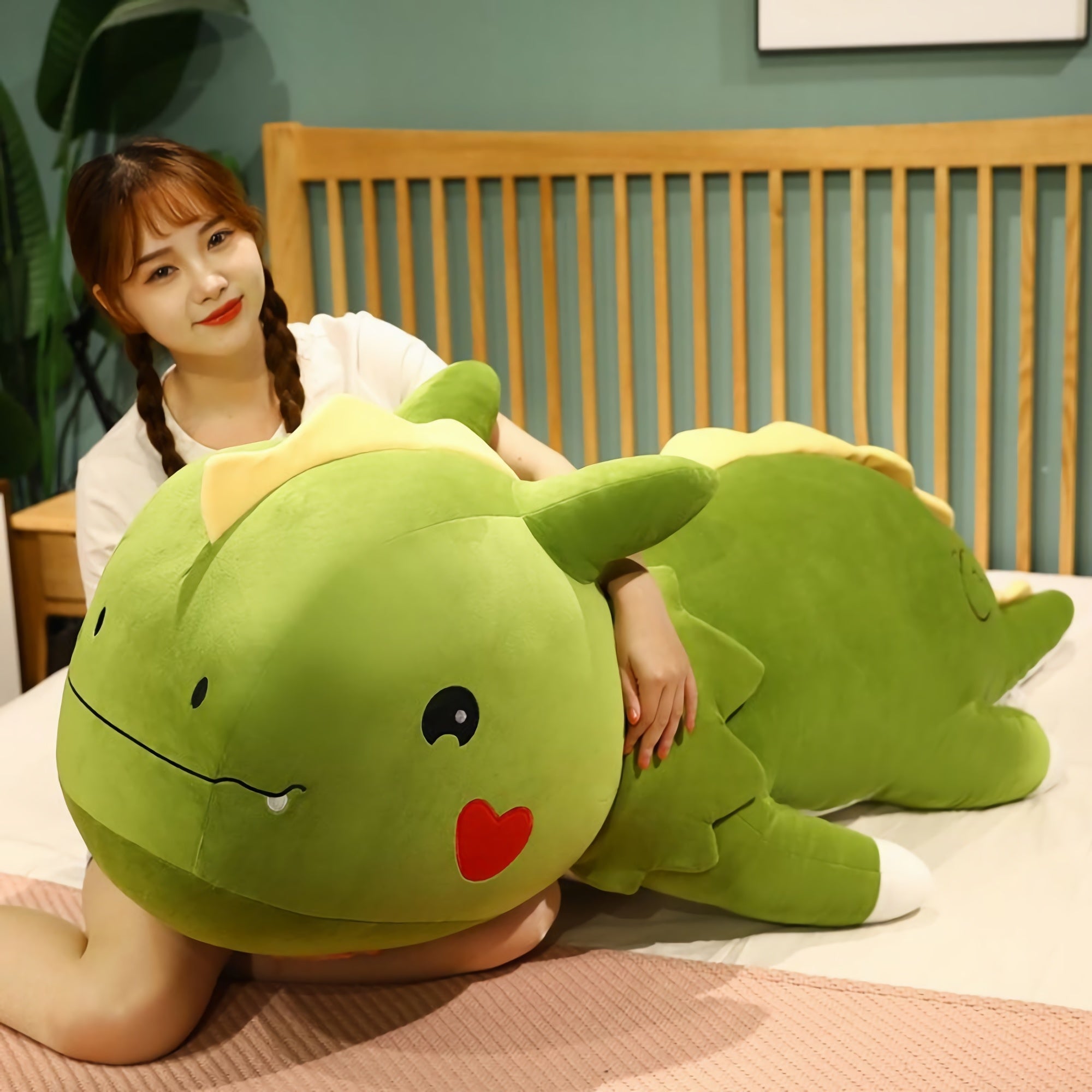 A person sits on a bed, hugging a large green dinosaur plush pillow with a smiling face and heart on its cheek.