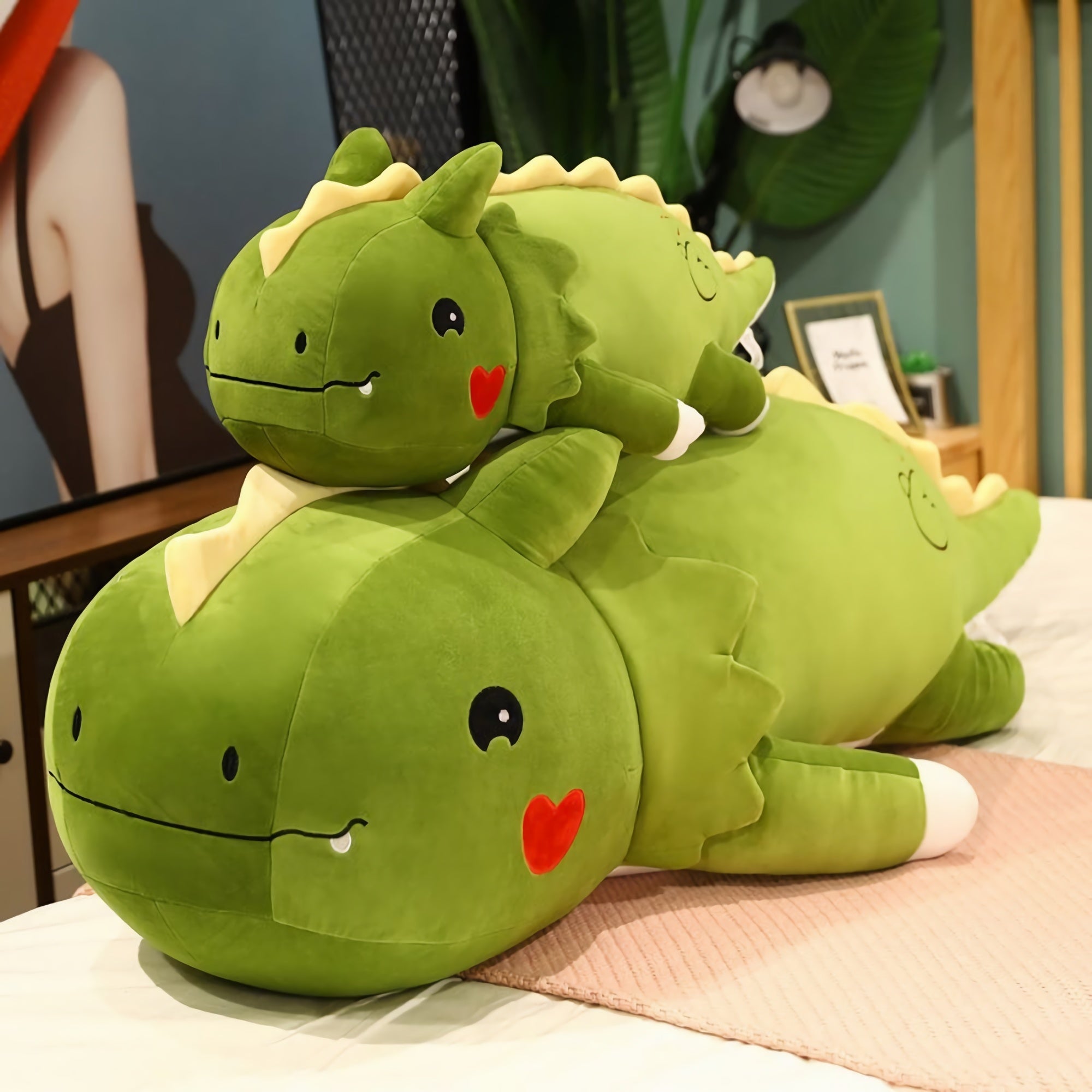 Green dinosaur plush pillows stacked on a bed, featuring a larger and a smaller version with cute facial expressions.