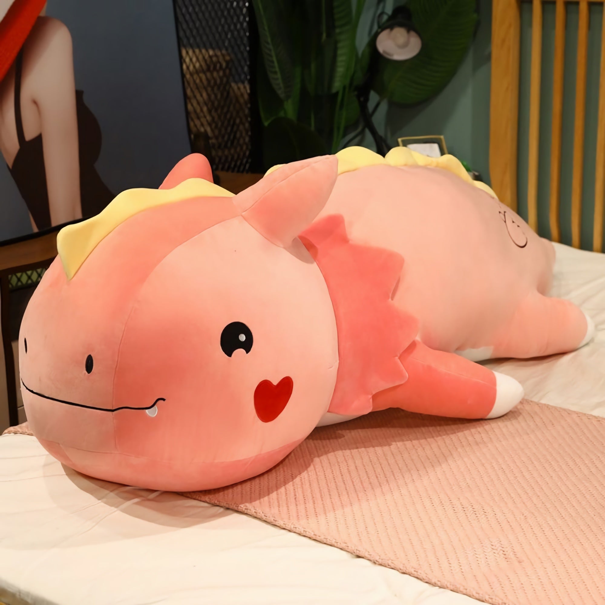 A large, pink dinosaur plush pillow with a smiling face and heart design, resting on a bed.