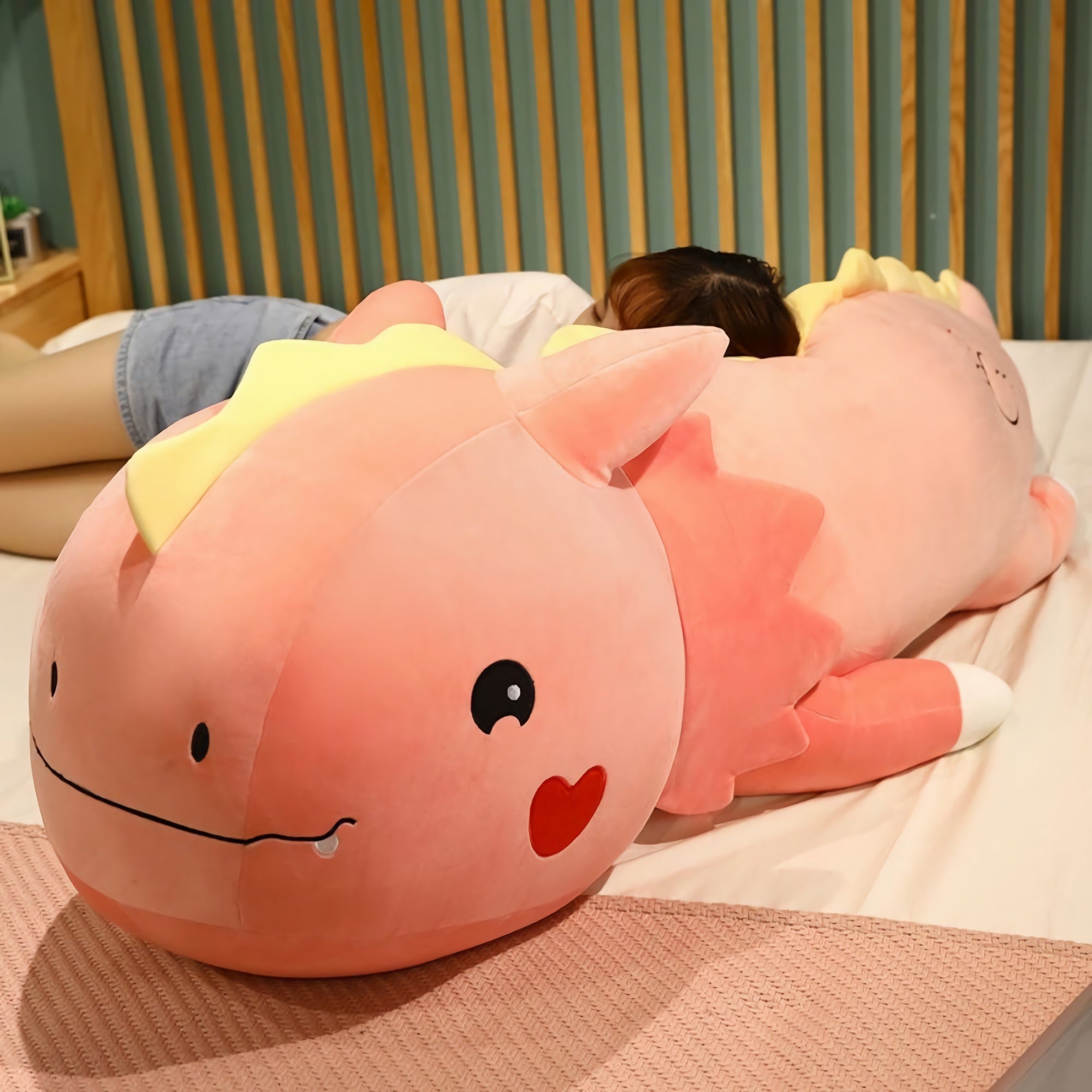 A large pink dinosaur plush pillow with a smiling face, resting on a bed beside a person lying down.