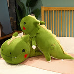 Green dinosaur plush pillow with a smiling face and heart detail, lying on a bed.