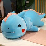 Blue dinosaur plush pillow with a smiling face and heart, lying on a bed with a pink mat.