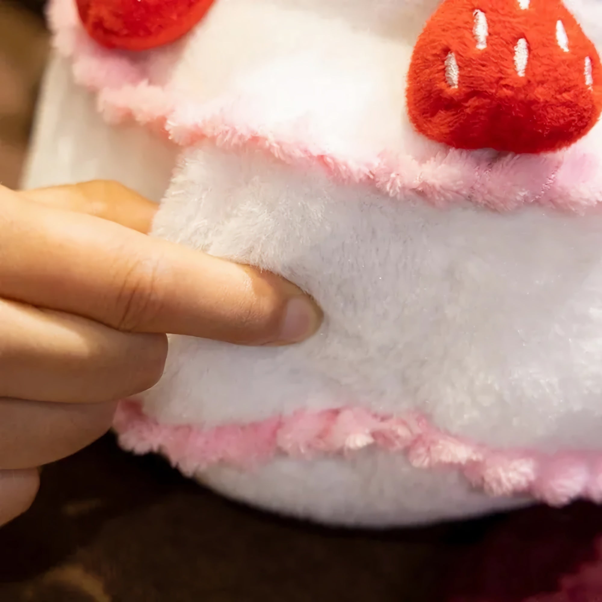 A hand pressing into a soft, plush pillow resembling a cake with pink trim and red strawberry details.