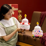 A person holds a plush birthday cake pillow with a candle, alongside another similar plush cake, in a cozy setting.