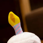Plush birthday cake pillow with a yellow candle and a striped pink and blue base.