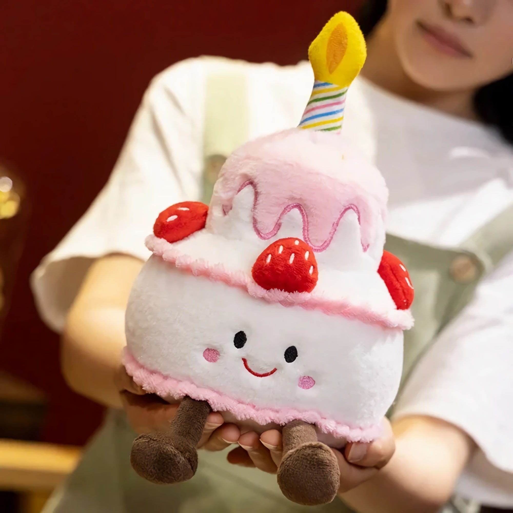 Plush pillow shaped like a birthday cake with a candle, strawberries, and a smiling face, held by a person.
