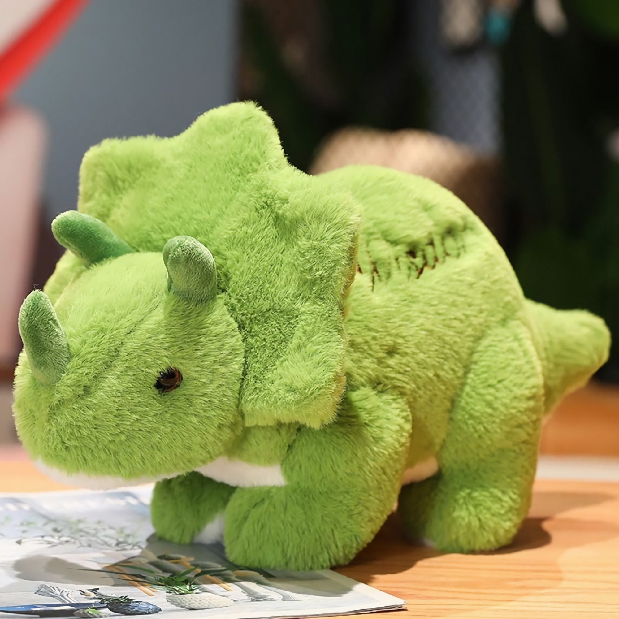 A green plush Triceratops toy with small horns and a frill, resting on a magazine on a wooden surface.