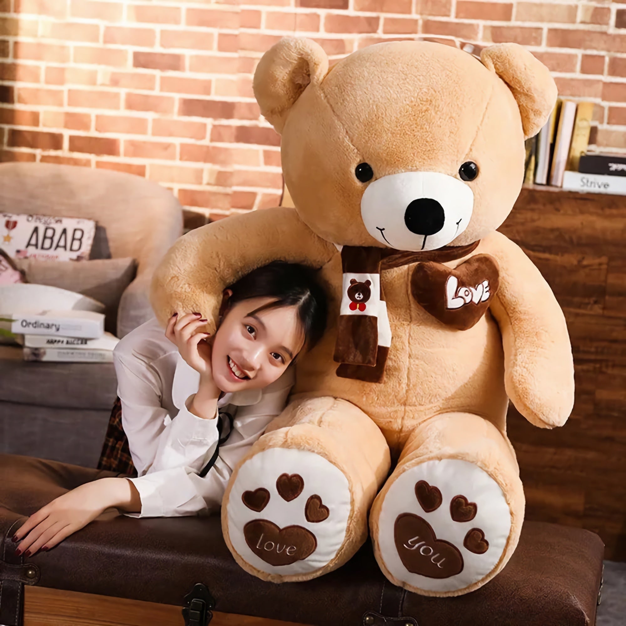 A girl smiles while hugging a large teddy bear with a scarf, sitting on a brown bench against a brick wall.
