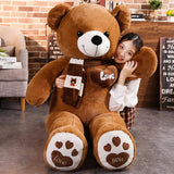 A girl hugs a large brown teddy bear plush with a scarf, sitting on a floor with a brick wall background.