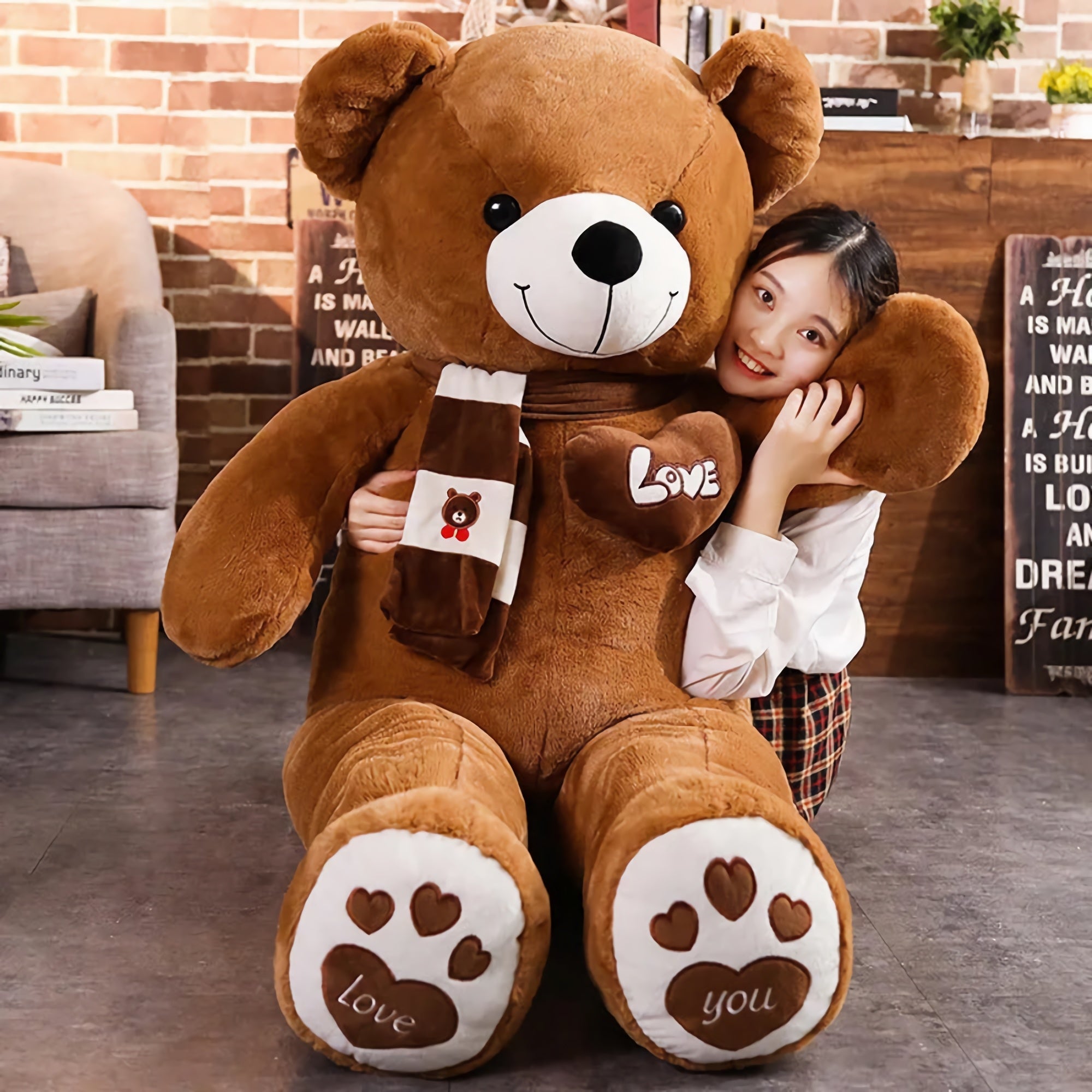 A girl hugs a large brown teddy bear plush with a scarf, sitting on a floor with a brick wall background.