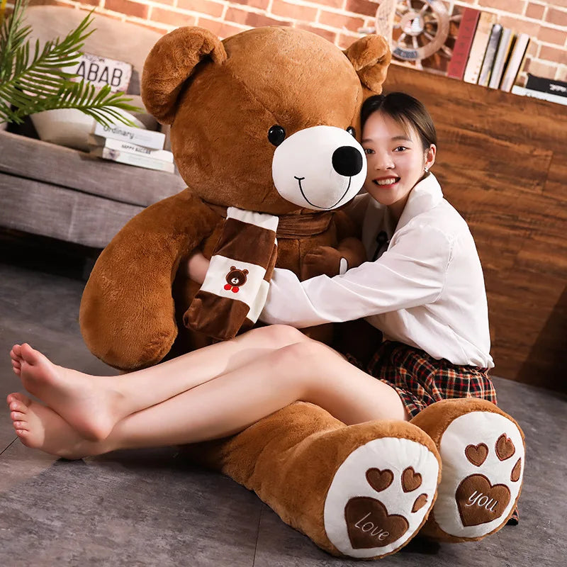 A girl sits beside a large brown teddy bear with a scarf, smiling and hugging it in a cozy room.